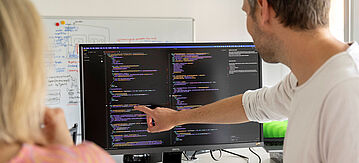 Eine männliche Person zeigt auf einen Monitor mit Computer-Code.