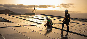 Bild von zwei Technikern, die an einer Photovoltaikanlage arbeiten, im Hintergrund eine Windkraftanlage