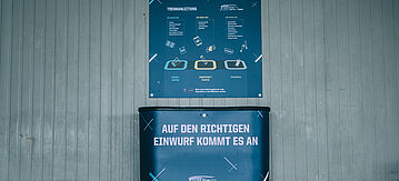 Blauer Abfallbehälter mit mehreren Abteilungen, darüber hängt ein Plakat mit den zugehörigen Abfallsorten. Auf dem Abfallbehälter steht: Auf den richtigen Einwurf kommt es an.