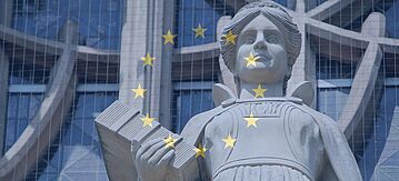 Statue einer Frau mit einem Gesetzbuch in der Hand, im Hintergrund ist die blaue Flagge der EU mit ihren gelben Sternen zu sehen.