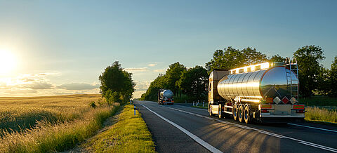 Lastwagen mit verchromten Tanks fahren bei Sonnenuntergang auf einer Asphaltstraße entlang des Maisfelds. Die Landschaft und die Straße spiegeln sich in einem silbernen Tankwagen.