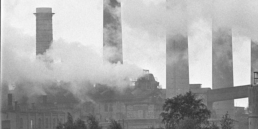 Schwarz-Weiß-Foto zeigt Fabrik-Gebäude und fünf Schornsteine, im Vordergrund Rauchwolken