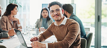 Junger Mann im Büro am Schreibtisch vorm Laptop schaut in die Kamera.