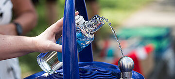 An einem Trinkwasserbrunnen wird eine Flasche mit Wasser gefüllt.. Weitere Informationen siehe Bildunterschrift