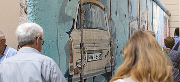 Besucherinnen und Bescuher vor einem Teil der Berliner Mauer mit einem Trabant-Auto drauf