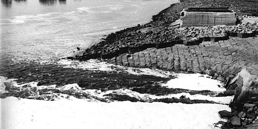 Schwarz-Weiß-Foto zeigt links einen Fluss und rechts das Ufer. Aus einem Rohr am Ufer fließen schäumende Abwässer in den Fluss.
