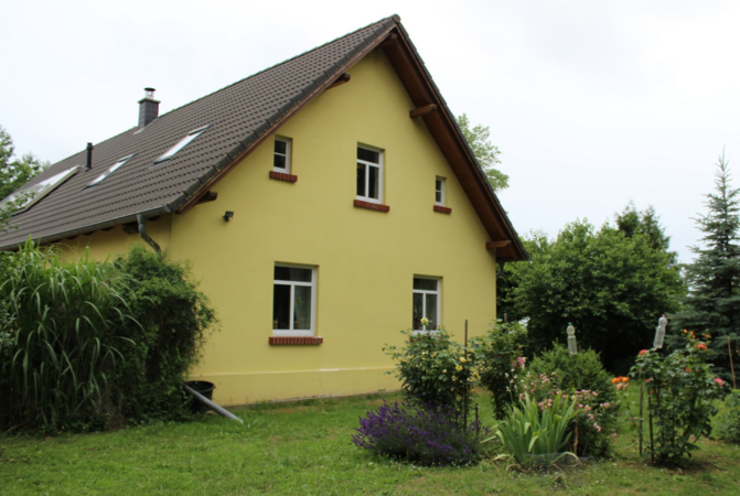 Ein Einfamilienhaus in Gelb steht inmitten von viel Grün auf einer Wiese. 