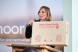 Eine Rednerin präsentiert Verpackungsmaterial, das unter anderem aus Pflanzen hergestellt wurde, die auf wiedervernässten Mooren gepflanzt wurden.
