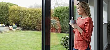 Eine Frau steht am Fenster mit einer Tasse und blickt in den Garten. 