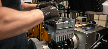 Eine Person mit schwarzen Arbeitshandschuhen setzt nach erfolgter Aufbereitung ein graues, geripptes Elektronikmodul in einer metallischen Vorrichtung an einer industriellen Montage-/Reparaturstation zusammen.