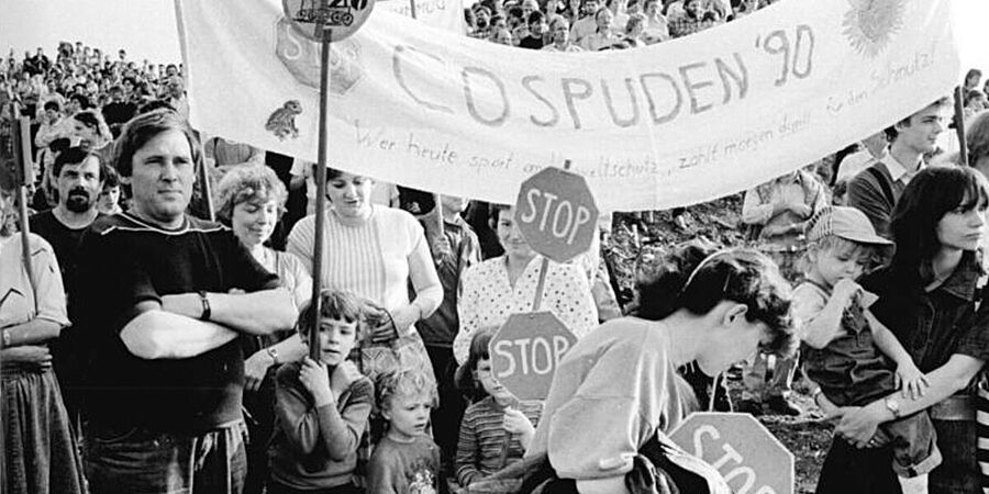Schwarz-Weiß-Foto einer Gruppe von Demonstrierenden. Mehrere Personen halten Stop-Schilder. Auf einem Transparent steht "Cospuden '90".