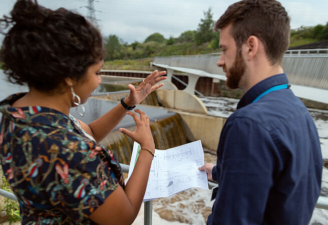 Zwei Umweltplaner*innen besprechen sich in der Nähe eines Staudammes. 
