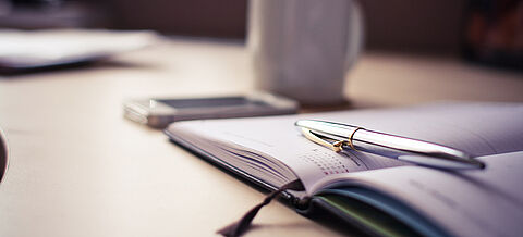 Silberner Kugelschreiber auf Terminplaner. Im Hintergrund sieht man eine Tasse und ein Mobiltelefon auf dem Tisch.