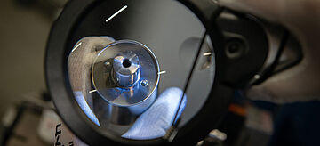 Zwei behandschuhte Hände inspizieren ein kleines Metallbauteil durch eine runde Lupenleuchte im Rahmen der Qualitätsprüfung nach erfolgter Reparatur/Remanufacturing.. Weitere Informationen siehe Bildunterschrift