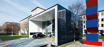 Bild des Effizienzhaus Plus von außen, davor steht ein Elektroauto, rechts daneben eine blaurote Skulptur