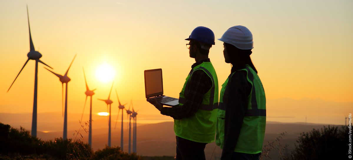 Zwei Arbeiter mit einem Laptop vor Windmühlen im Sonnenuntergang